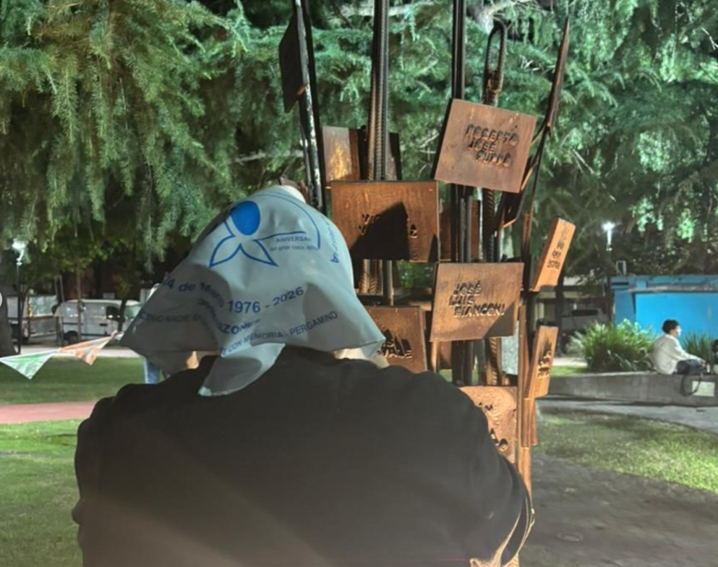 Una persona con pañuelo blanco en la cabeza frente al memorial de la plaza.
