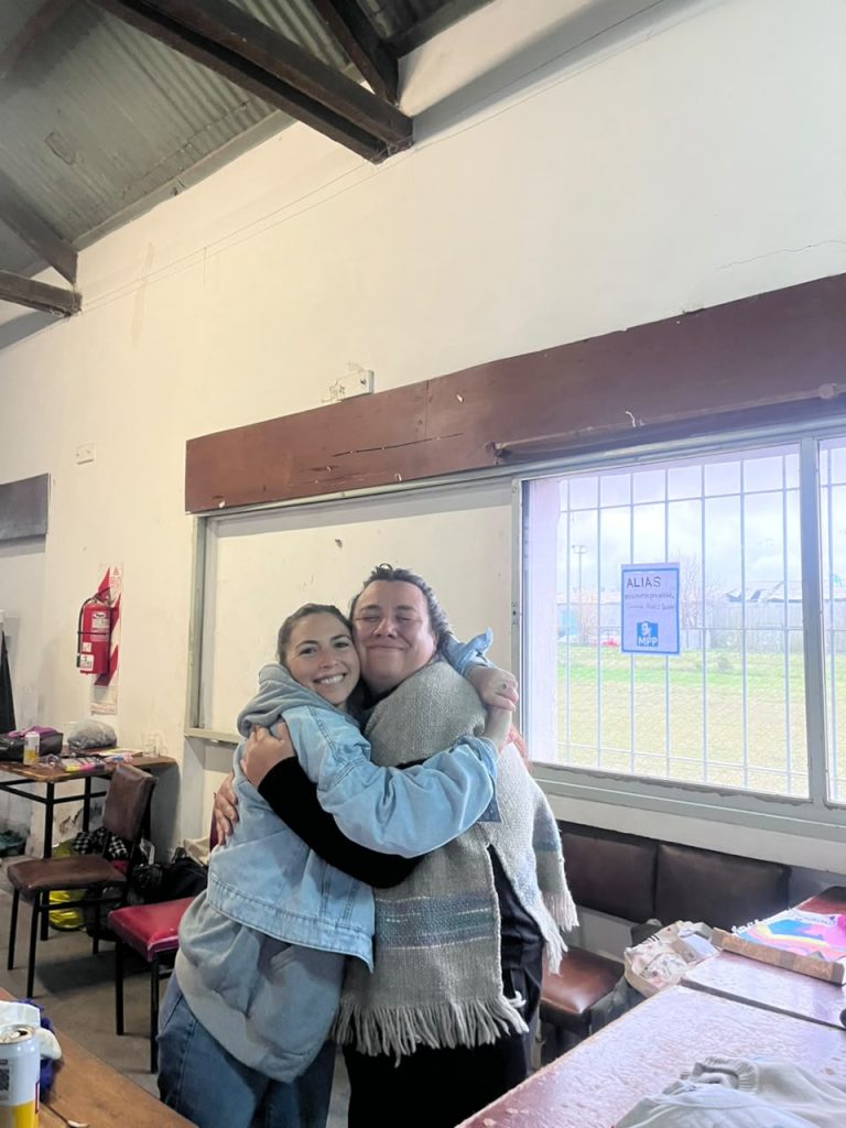 Dos compañeras se abrazan en la peña, junto a la mesa de la feria.