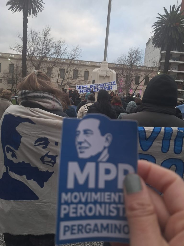 Un volante del Movimiento Peronista Pergamino en primer plano, con la movilización de fondo.